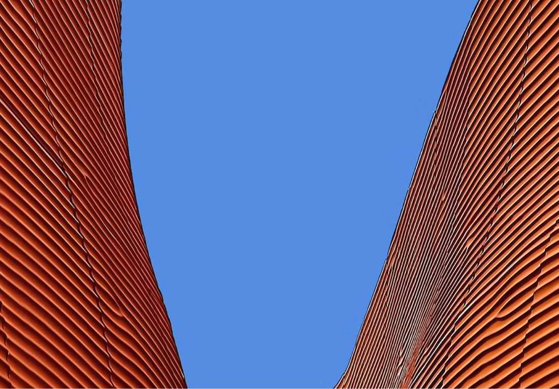 Close-up view of a modern architectural structure with curved, rust-colored metal panels against a clear blue sky. The panels have a ribbed texture and create an undulating form, reminiscent of waves or sand dunes.