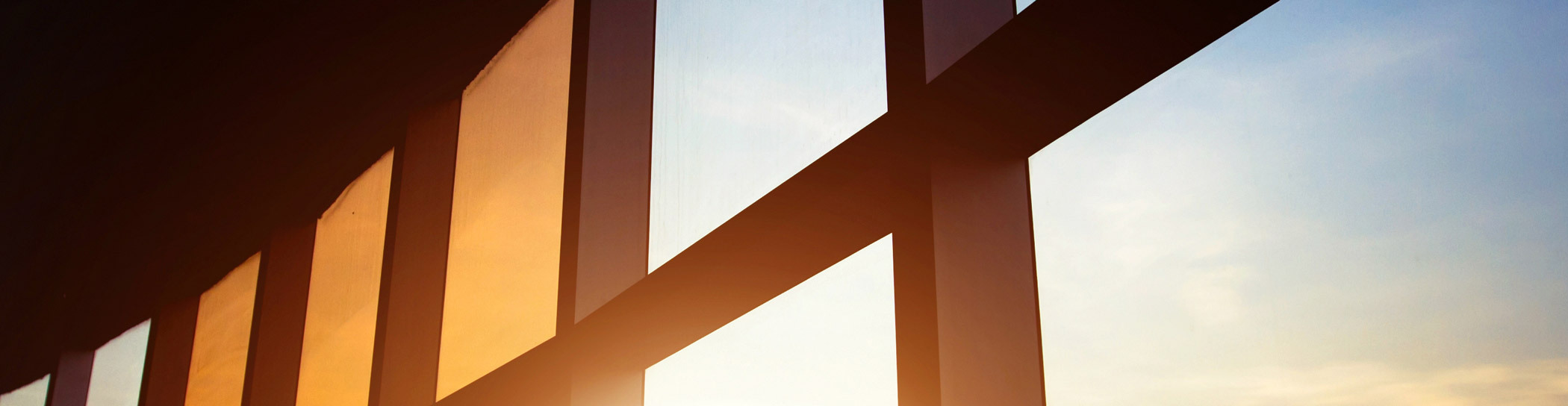 A photograph of a sunset viewed through the silhouette of a geometric window structure, with the sun casting warm hues across the sky and creating a contrast between the dark window frame and the illuminated outside.