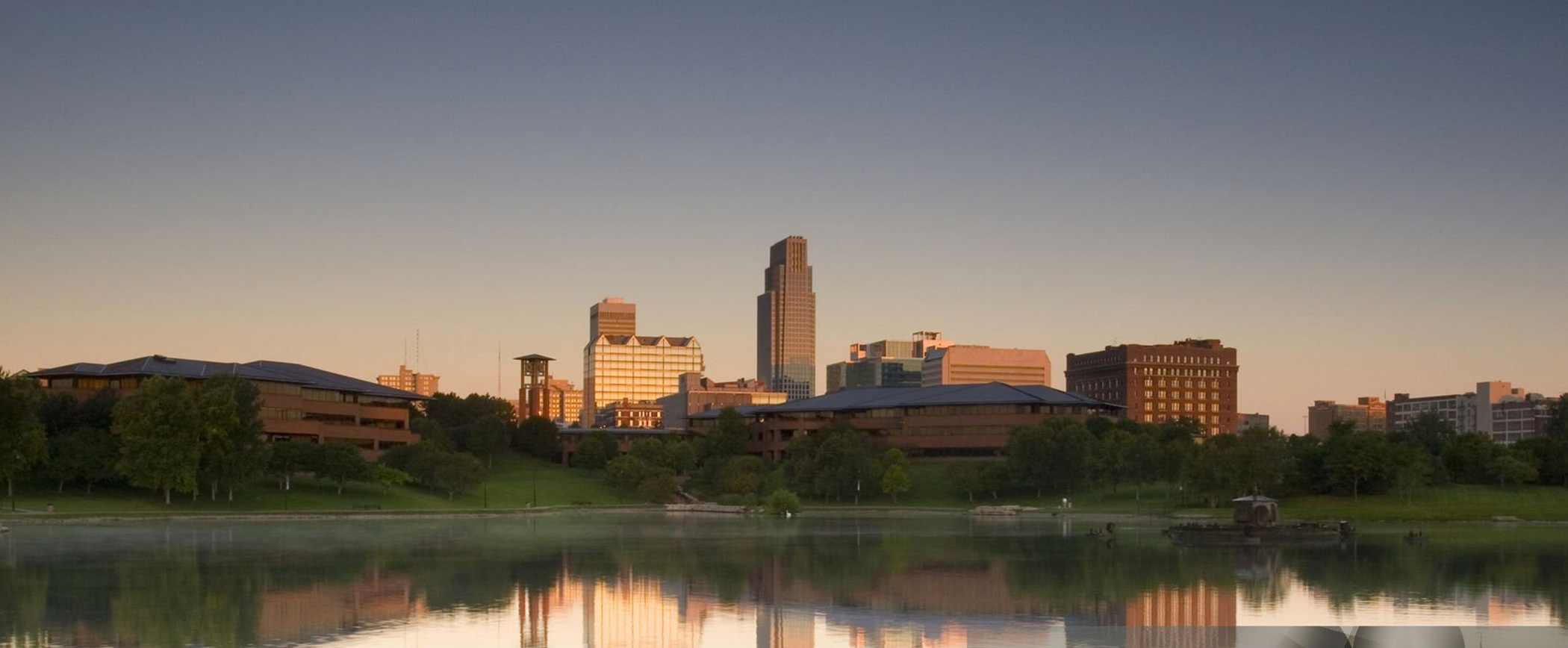 A panoramic view of downtown Omaha at dusk, featuring a serene lake in the foreground with reflections of the skyline. The cityscape includes various buildings with different architectural styles and heights, with the tallest building standing prominently