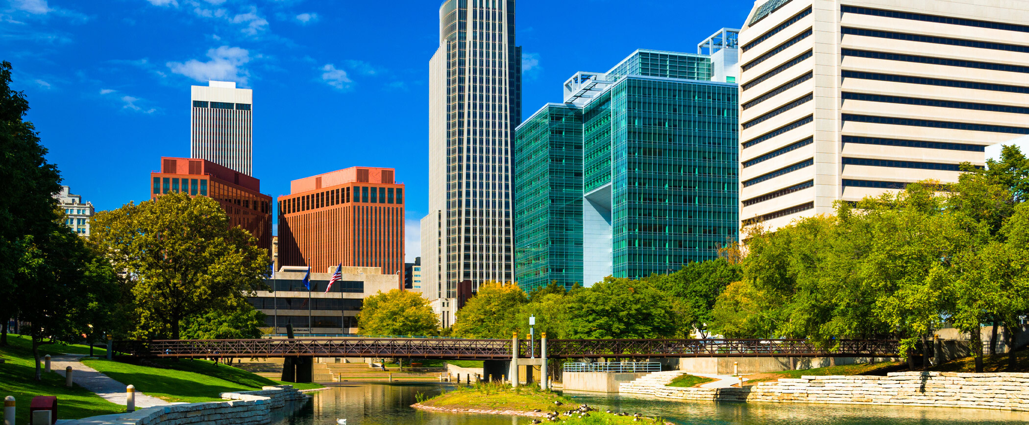 Downtown Omaha skyline with modern office buildings, a clear blue sky, and a calm river reflecting the cityscape and greenery.