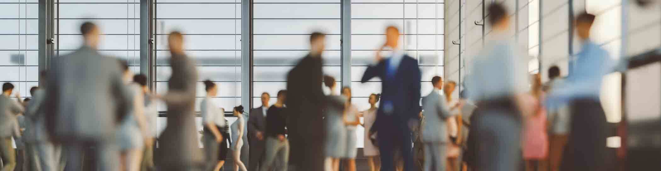 A blurred image of a busy indoor gathering with numerous people dressed in business attire, engaging in conversations and networking. The background features large windows allowing natural light to fill the space, creating a modern and professional enviro
