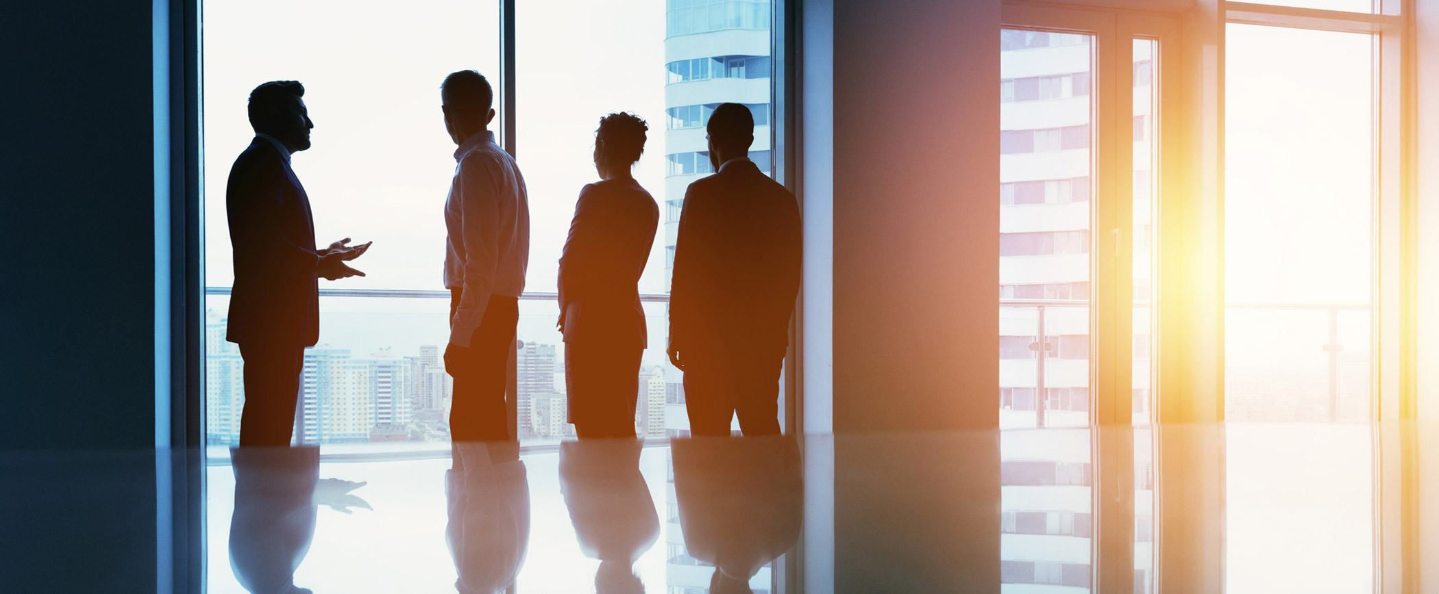 Silhouettes of four business professionals standing and conversing in front of large windows in a modern office setting. The cityscape is visible through the windows, and sunlight is streaming into the room, creating a bright and professional atmosphere.