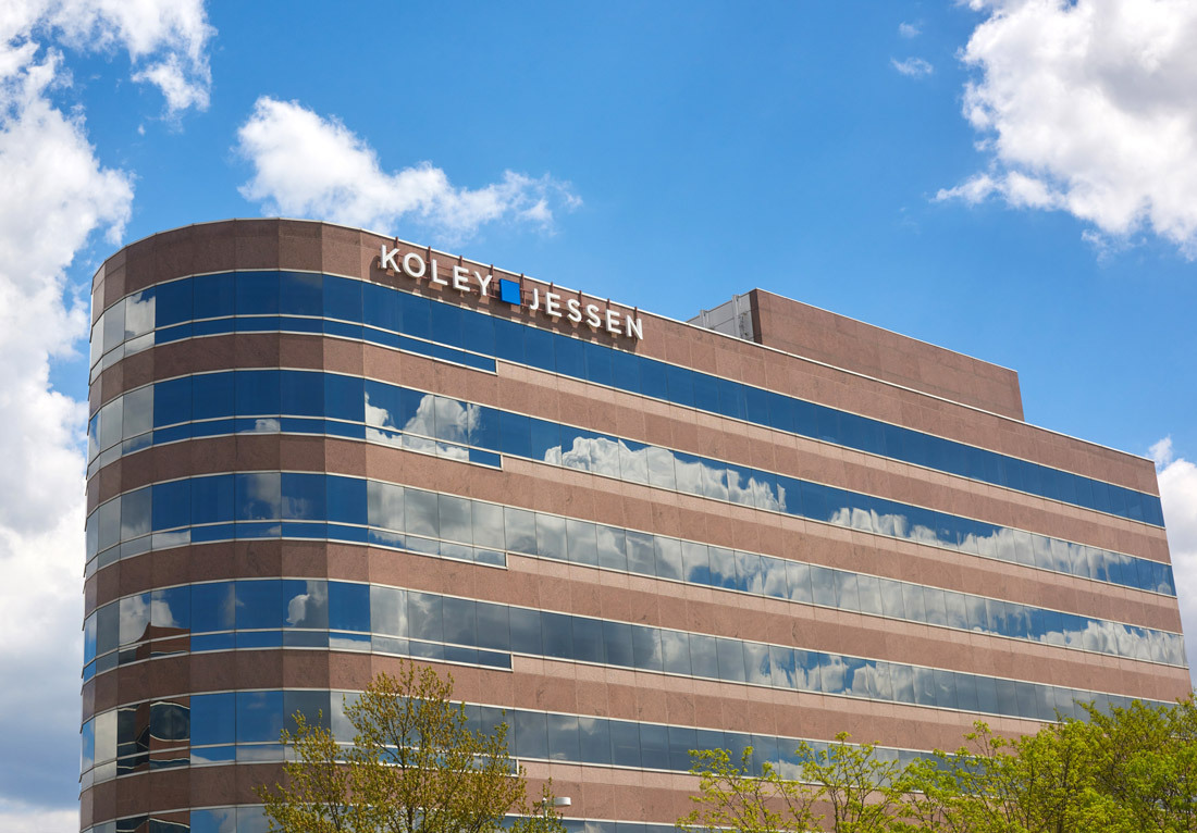 A modern office building with a curved facade, featuring large reflective windows. The name "Koley Jessen" is prominently displayed at the top in white letters against the brown and glass exterior. The building is set against a bright blue sky with scatte