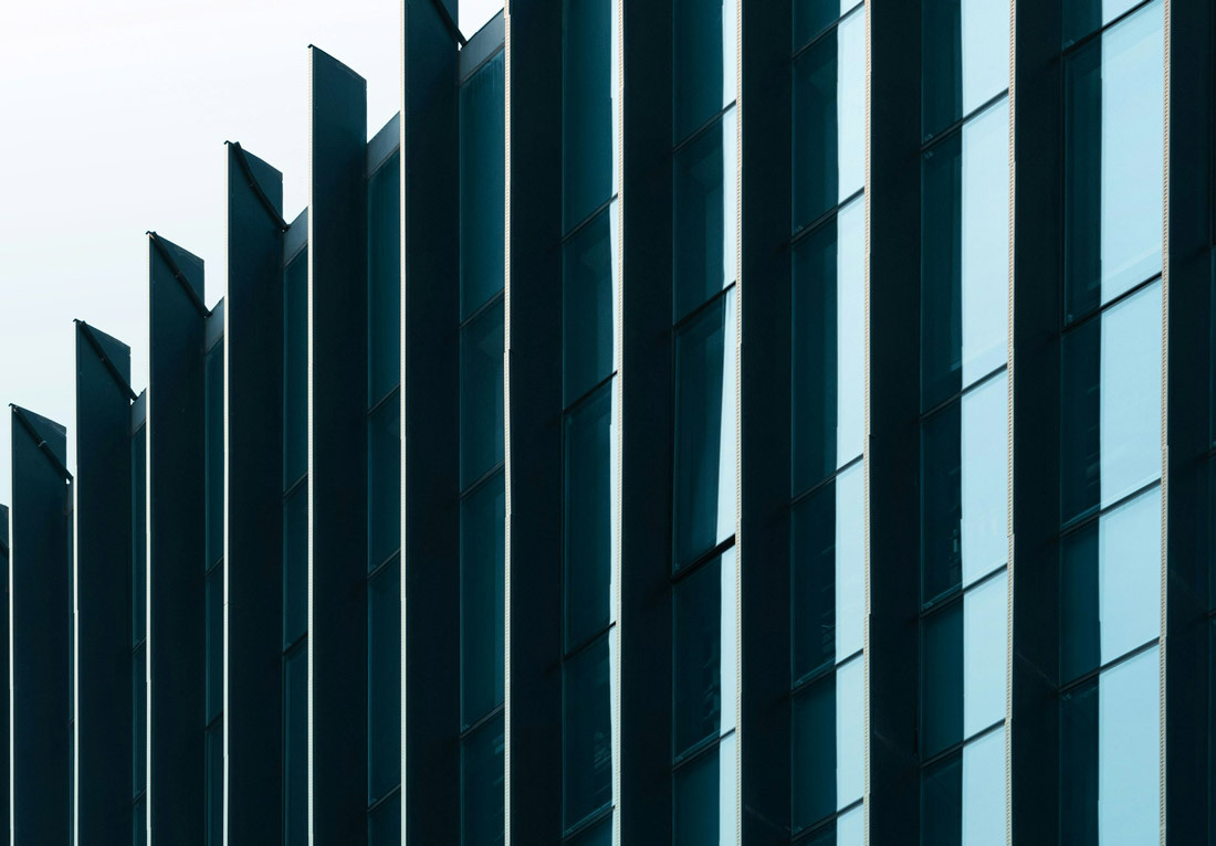 Close-up view of a modern building facade featuring vertical, evenly spaced, dark-colored fins and large glass windows reflecting the sky.