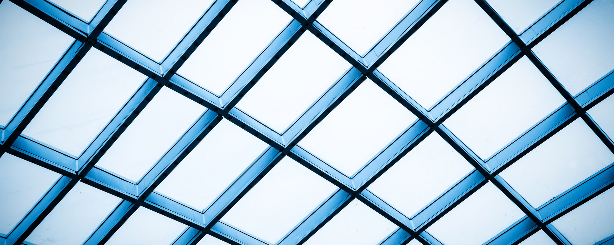 A view of a geometric glass ceiling made up of diamond-shaped panels framed by blue metal supports. The panels are arranged in a grid pattern, allowing light to filter through, creating a bright and airy atmosphere.