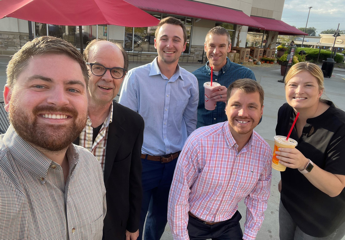 A group of six smiling people, three men and three women, are standing outdoors in front of a building with red umbrellas. They appear to be enjoying a casual outing, with three of them holding beverages with red straws. They are dressed in business casua