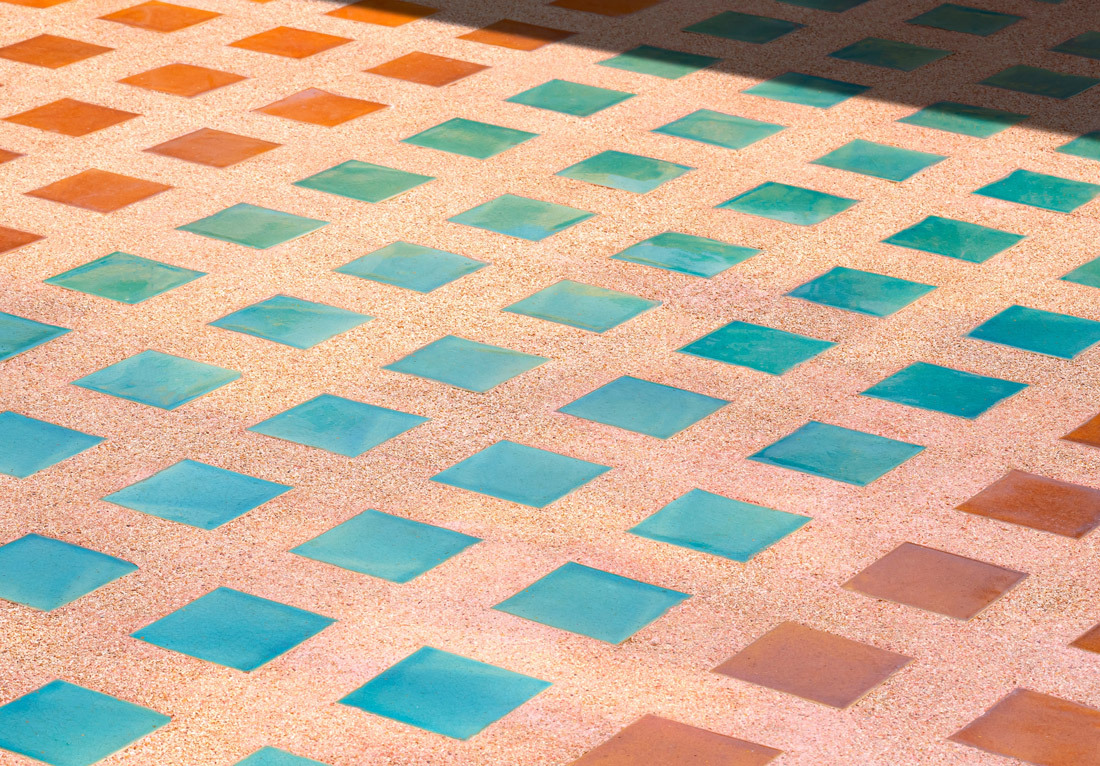 A close-up view of a floor with a geometric pattern of square tiles in shades of orange and blue, illuminated by warm sunlight.