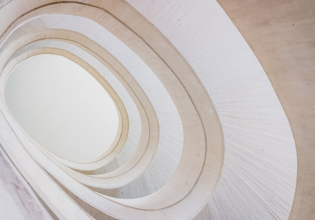 An upward view of an architectural structure featuring multiple layers of curved, beige-colored concrete forms, creating an oval-shaped opening at the top. The light from above softly illuminates the smooth, concentric curves, highlighting the texture of