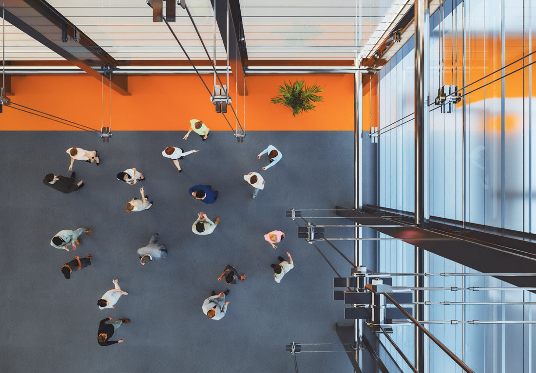 Overhead view of a modern office space with individuals walking and interacting. The floor is gray, and there are orange accents on the walls. Large windows allow natural light to enter, and a staircase with metal railings is visible on the left. A potted