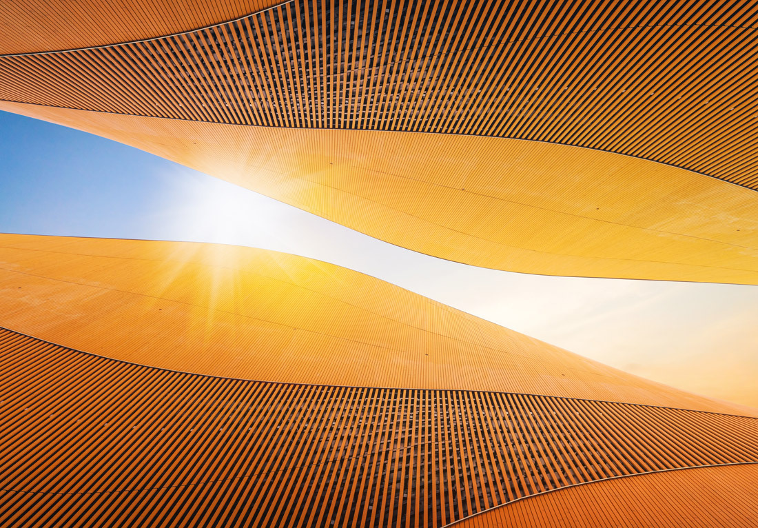 A series of curved orange and yellow bands with intricate line patterns against a blue sky, creating an abstract design that resembles sand dunes under a sunset or sunrise. The sun is positioned at the upper center where the bands converge, casting light
