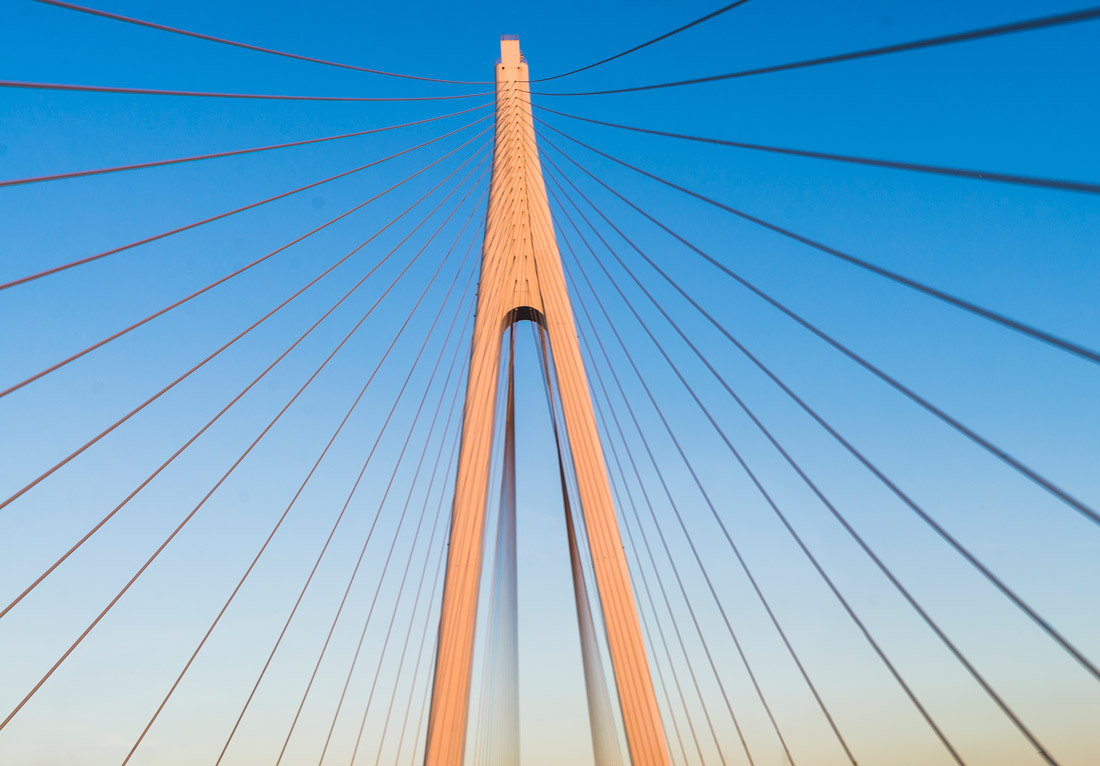 Close-up view of a modern suspension bridge against a clear blue sky. The bridge features a tall, slender pylon with numerous cables fanning out symmetrically from the top of the pylon to the deck below. The sunlight illuminates the orange-hued structure,