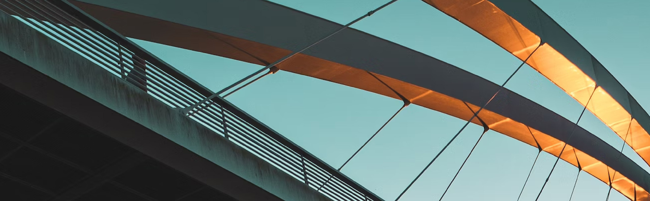 A close-up view of a modern bridge against a clear sky. The bridge features a sleek, curved design with an underside illuminated by warm sunlight, creating a contrast of light and shadow. The railing and cables are visible, adding to the architectural det