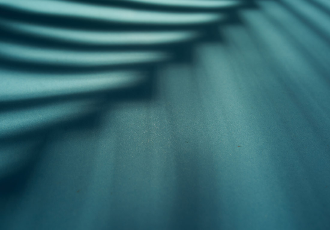 Close-up photograph of a smooth teal surface with undulating shadows creating a pattern of curved lines and soft light gradients, suggesting a tranquil and abstract aesthetic.