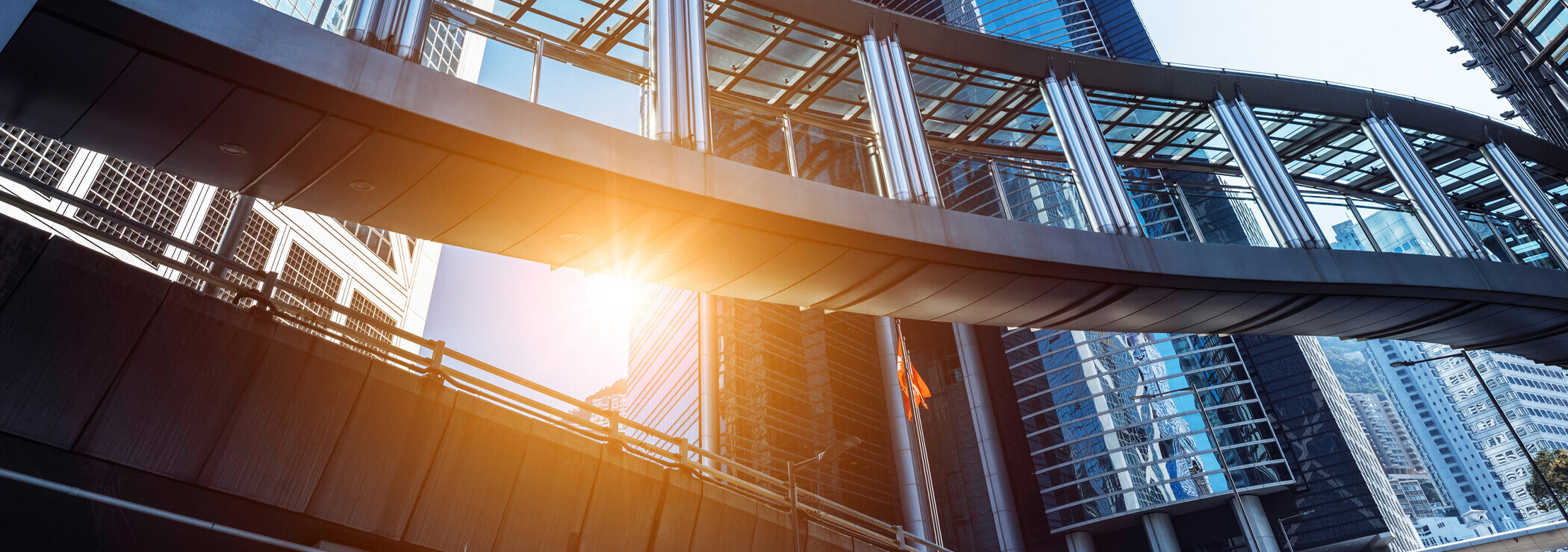 A modern pedestrian bridge with a metal and glass structure, connecting two buildings against a backdrop of skyscrapers under a clear blue sky. The sun is visible and creates lens flare, adding to the visual interest of the scene.