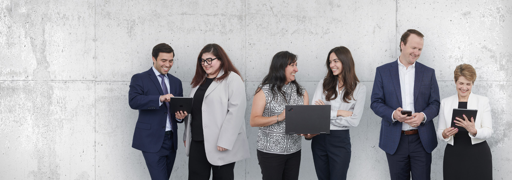 Group of six professionals from Koley Jessen standing against a textured gray wall, dressed in business attire. Two individuals hold a tablet, another holds a laptop, and one is using a smartphone.