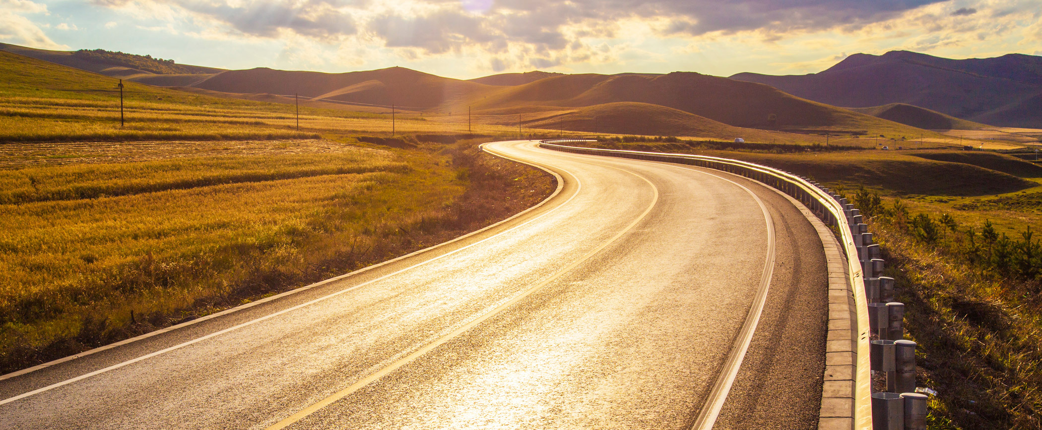 A panoramic view of a winding road stretching through a hilly landscape during golden hour, with the sun casting a warm glow on the asphalt and surrounding grassy fields, leading towards distant mountains under a partly cloudy sky.