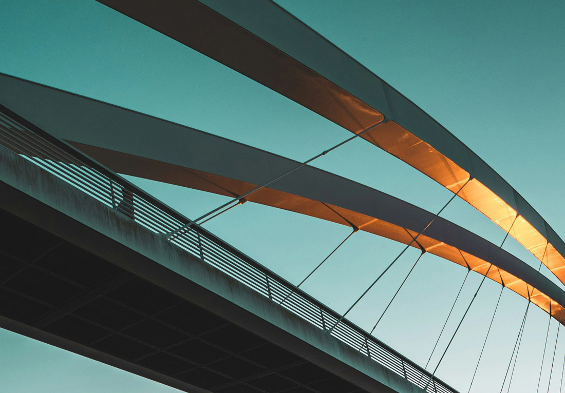 A close-up view of a modern bridge against a clear sky. The bridge features a sleek, curved design with an underside illuminated by warm sunlight, creating a contrast of light and shadow. The railing and cables are visible, adding to the architectural det